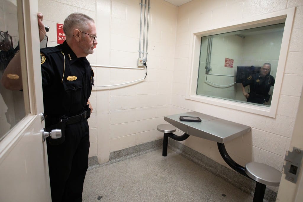 Sheriff Richard Fuller holds the door open to a small room with beige stone walls, a large plate glass window, and a metal table with two attached stools.