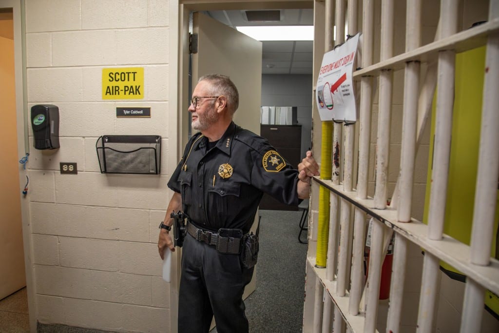 Sheriff Richard Fuller holds open a steel-barred jail cell door. The bars are painted beige but the paint is visibly worn and peeling.